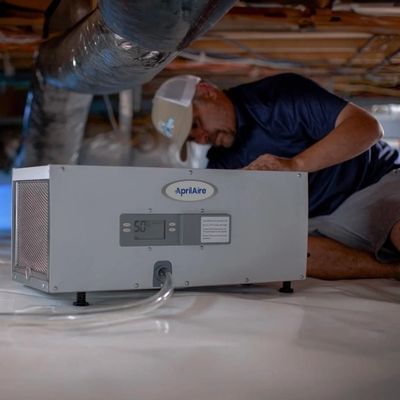 Technician installing an Aprilaire air purifier in a crawlspace beneath a house