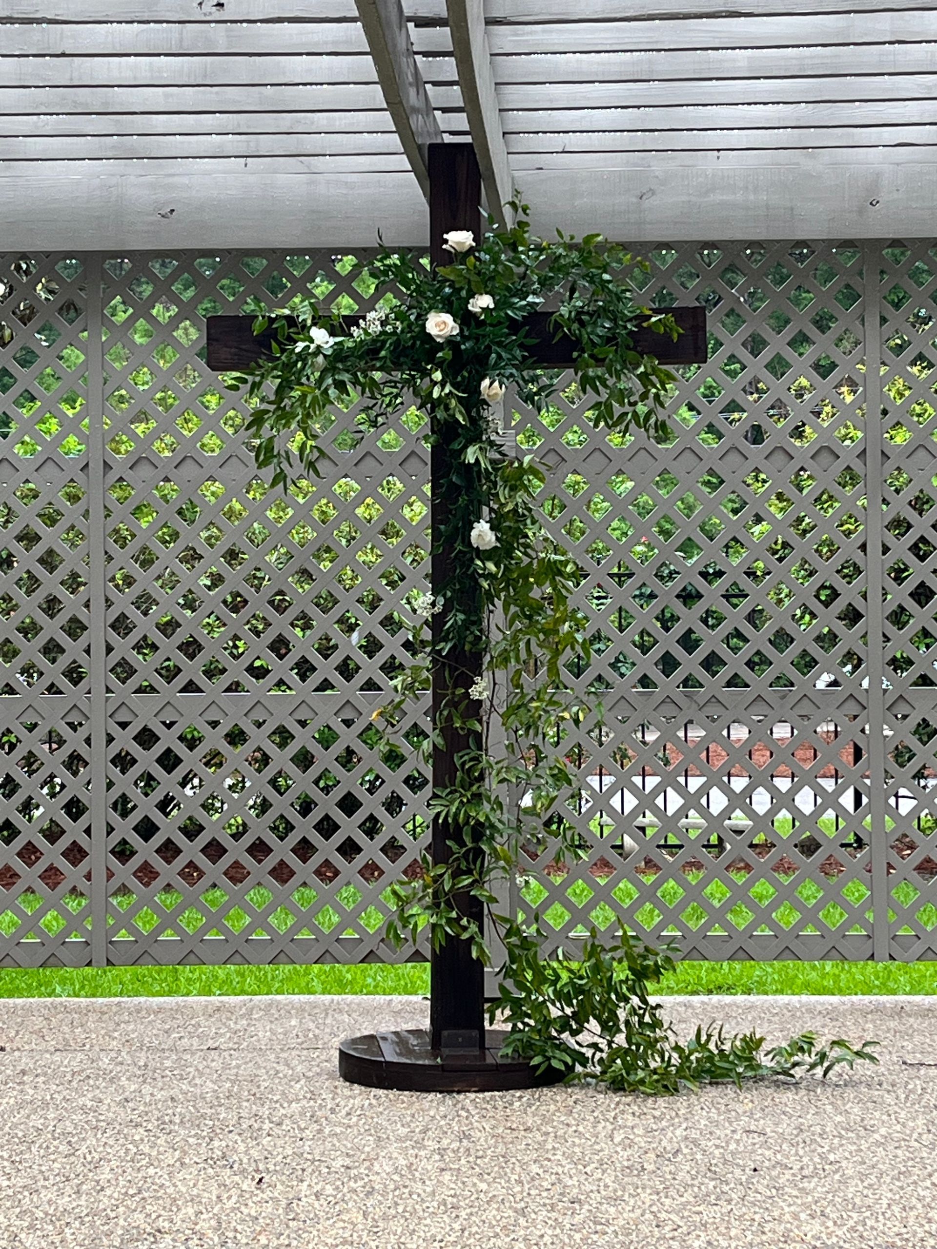 A cross decorated with flowers and greenery is sitting in front of a lattice fence.