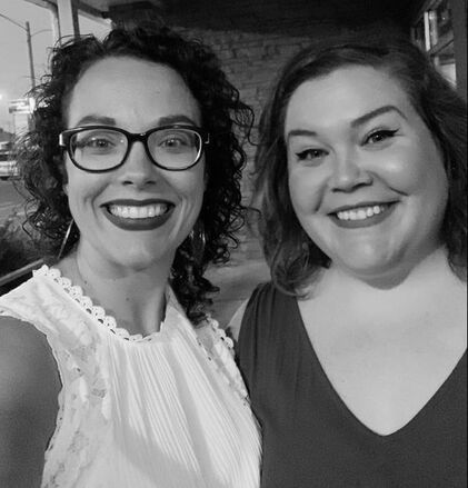 Two smiling people posing closely together indoors, one in glasses and a white blouse.