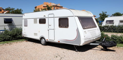 White camper trailer parked on a gravel lot under a sunny sky, with trees and buildings in the background