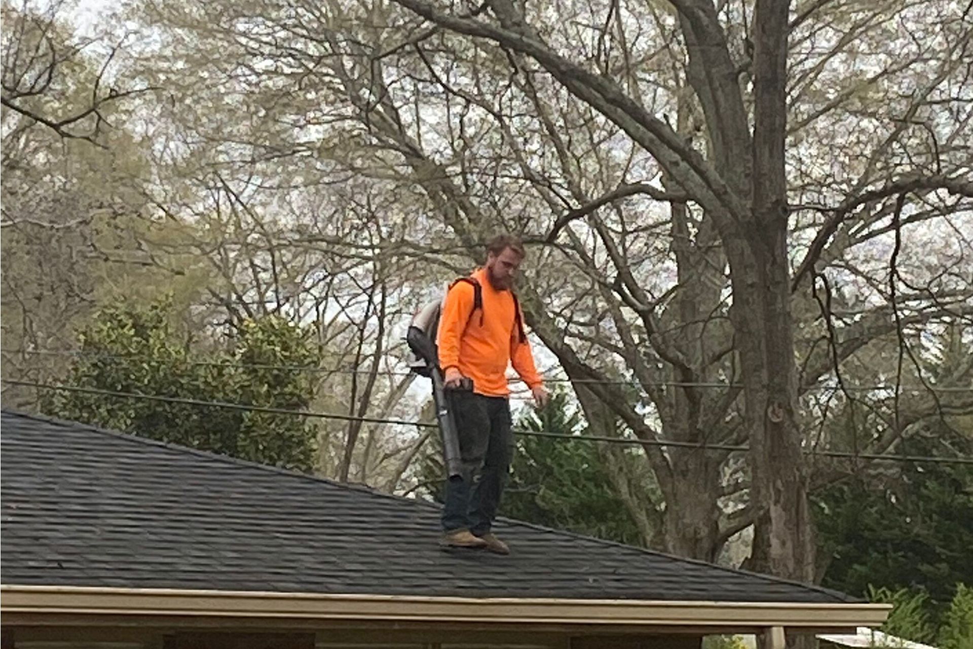 Person in an orange safety vest standing on a shingled roof near trees.