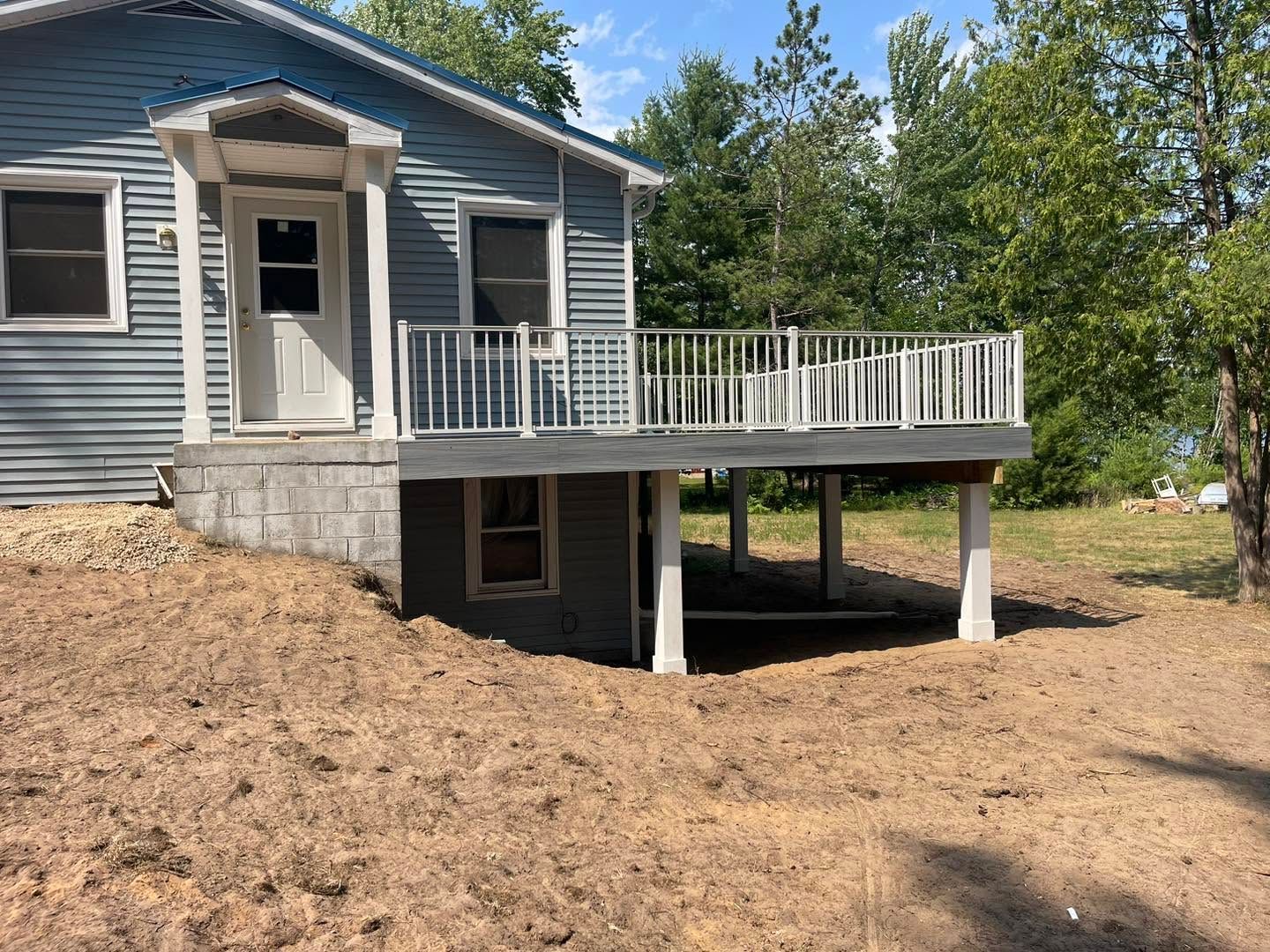 Gray house with a raised white porch and deck over a dirt yard, viewed from the side.