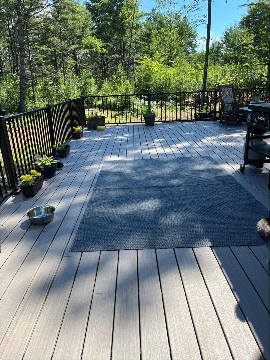 Sunlit wooden deck with black railing, potted plants, and outdoor furniture overlooking trees