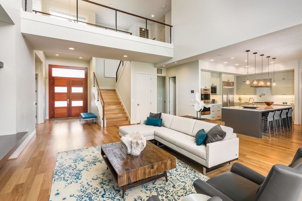 Bright open-concept living room with white sofa, staircase, loft railing, and kitchen/dining area.