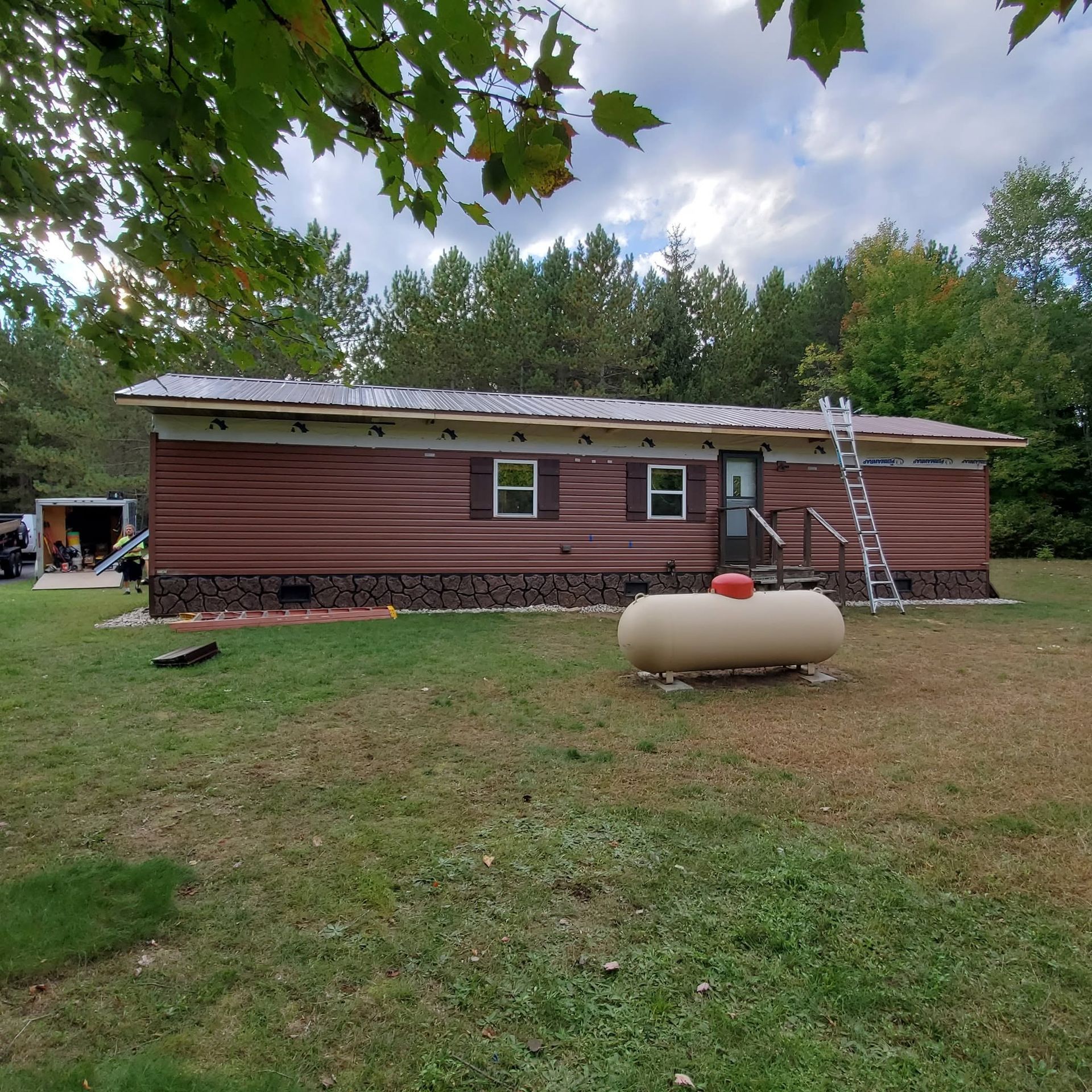Single-story brown house with a white ladder and propane tank in a grassy yard, surrounded by trees.