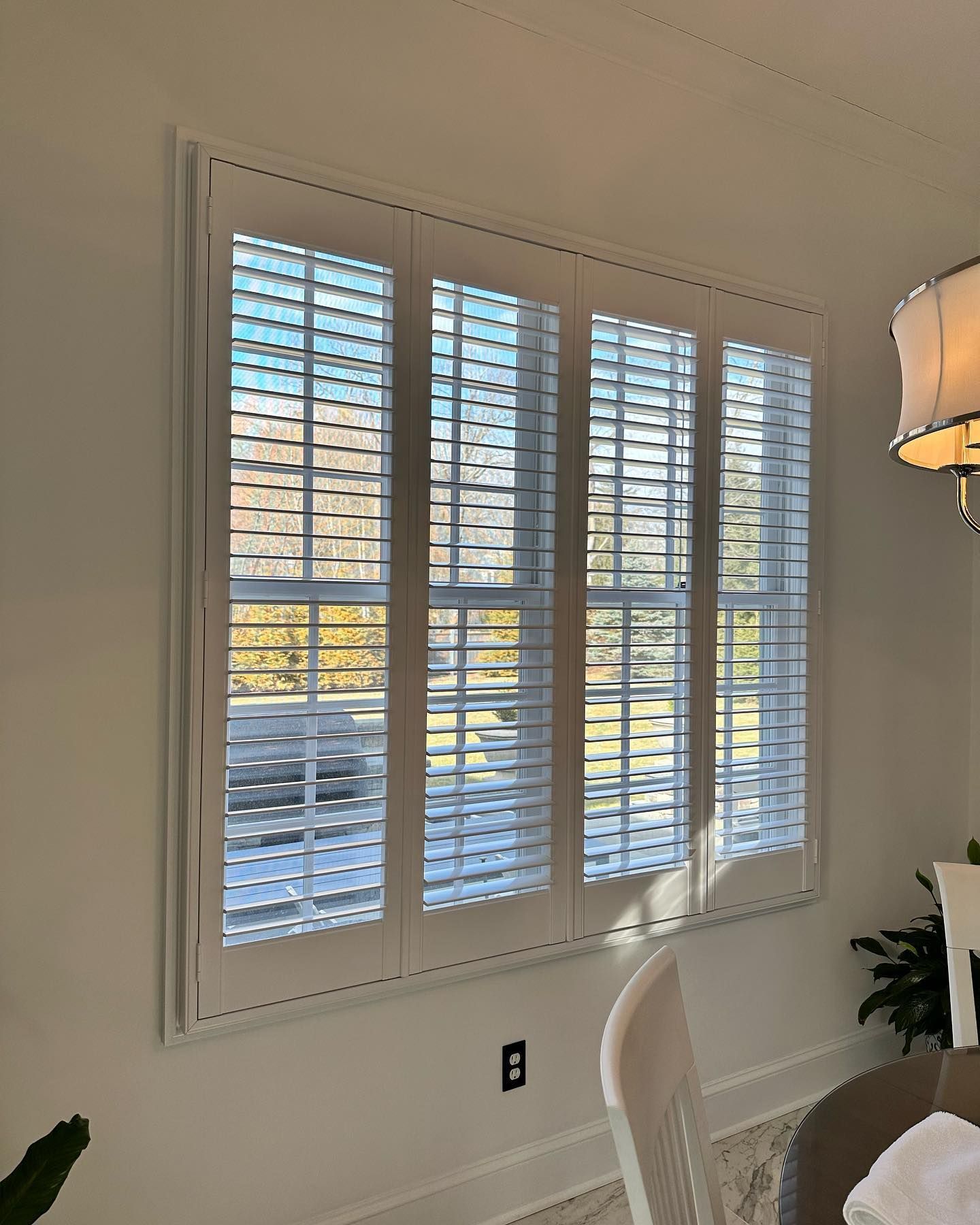 White window shutters in a room with a partial view of outdoor trees and a table with a chair.