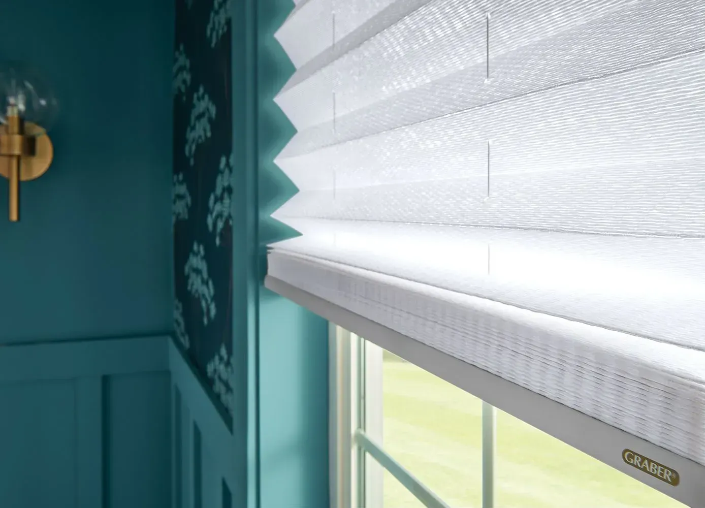 White pleated window shade in front of a teal wall and window.