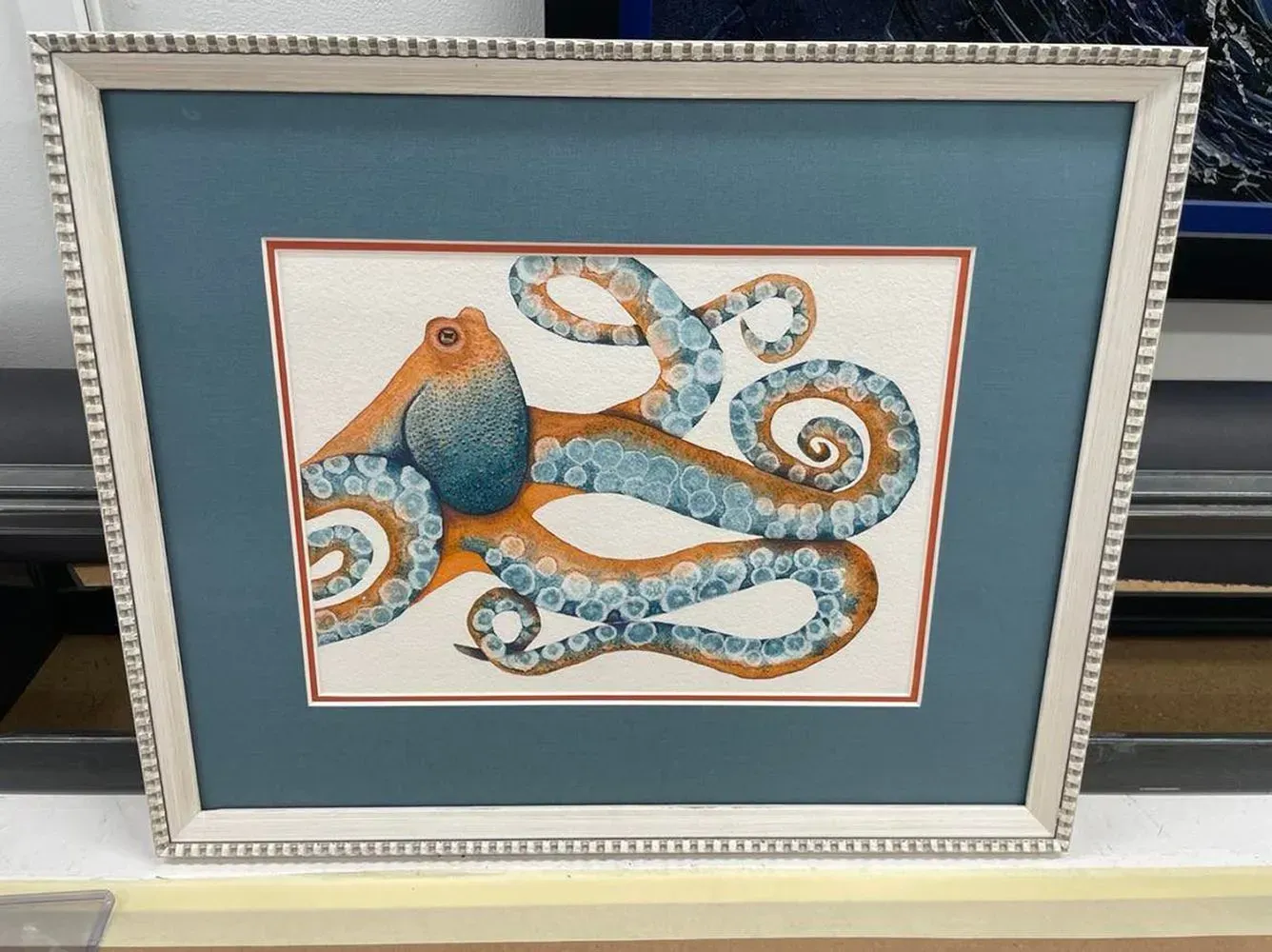 Framed artwork of an orange and blue octopus on white, set against a teal background.