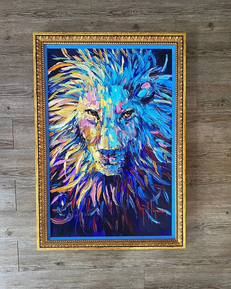 Lion portrait in vibrant blue, yellow, and purple oil paint, framed in gold against a wood background.