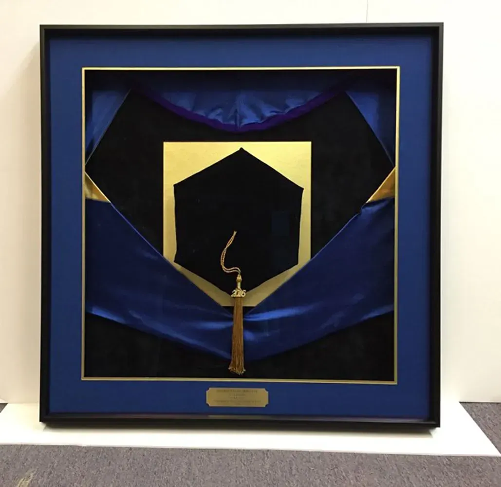 Framed display of a black graduation cap with gold tassel and blue and black academic regalia.