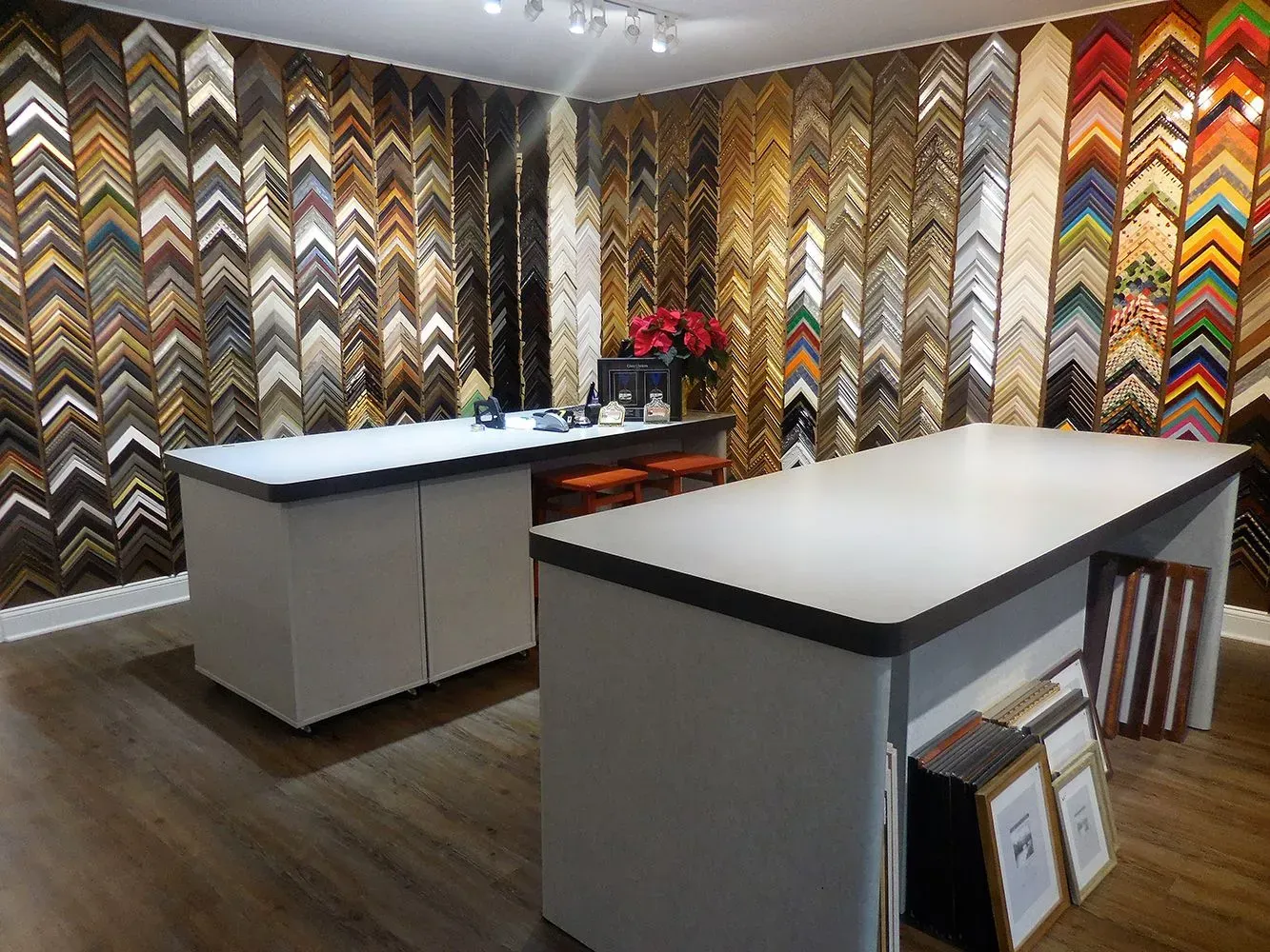 Frame shop interior with a variety of frames on display, two white work tables, and hardwood flooring.