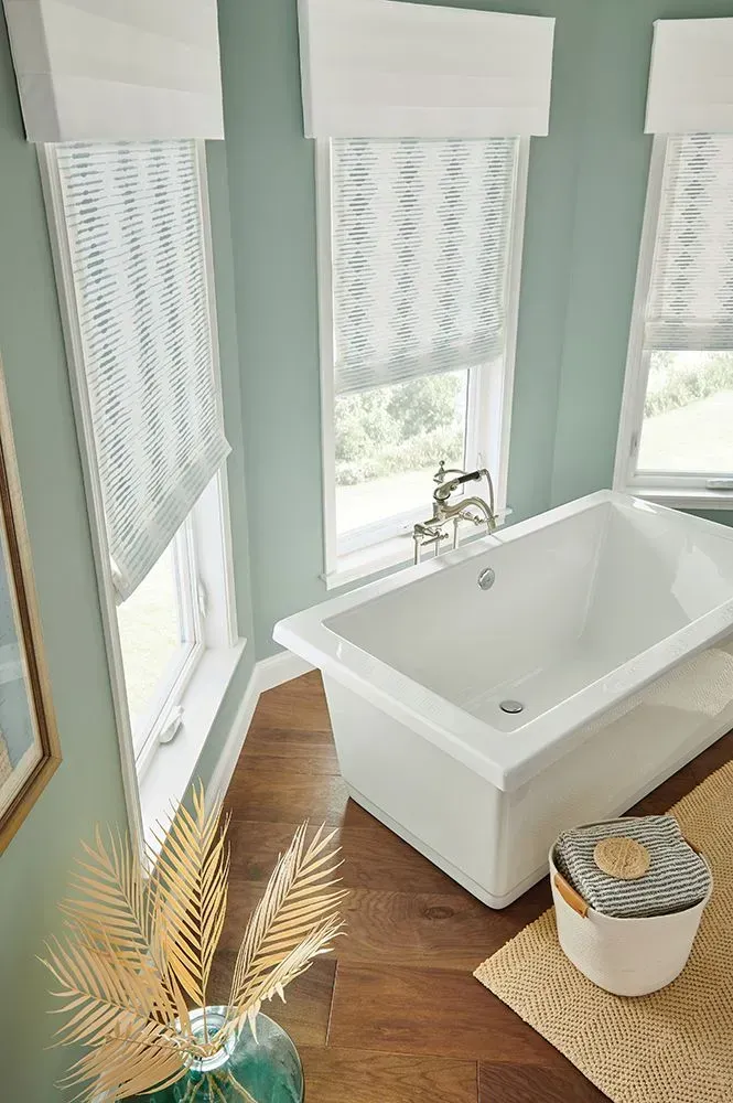 Bathroom with a white soaking tub, three windows with shades, and a wooden floor.