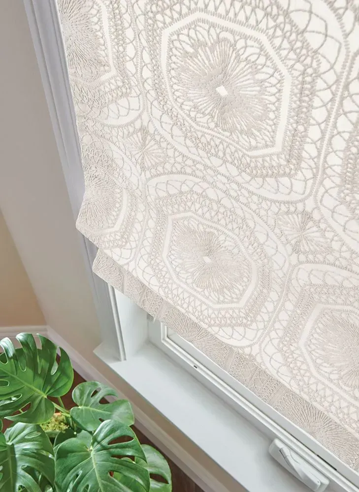 Beige patterned Roman shade in a white window frame, next to a Monstera plant.