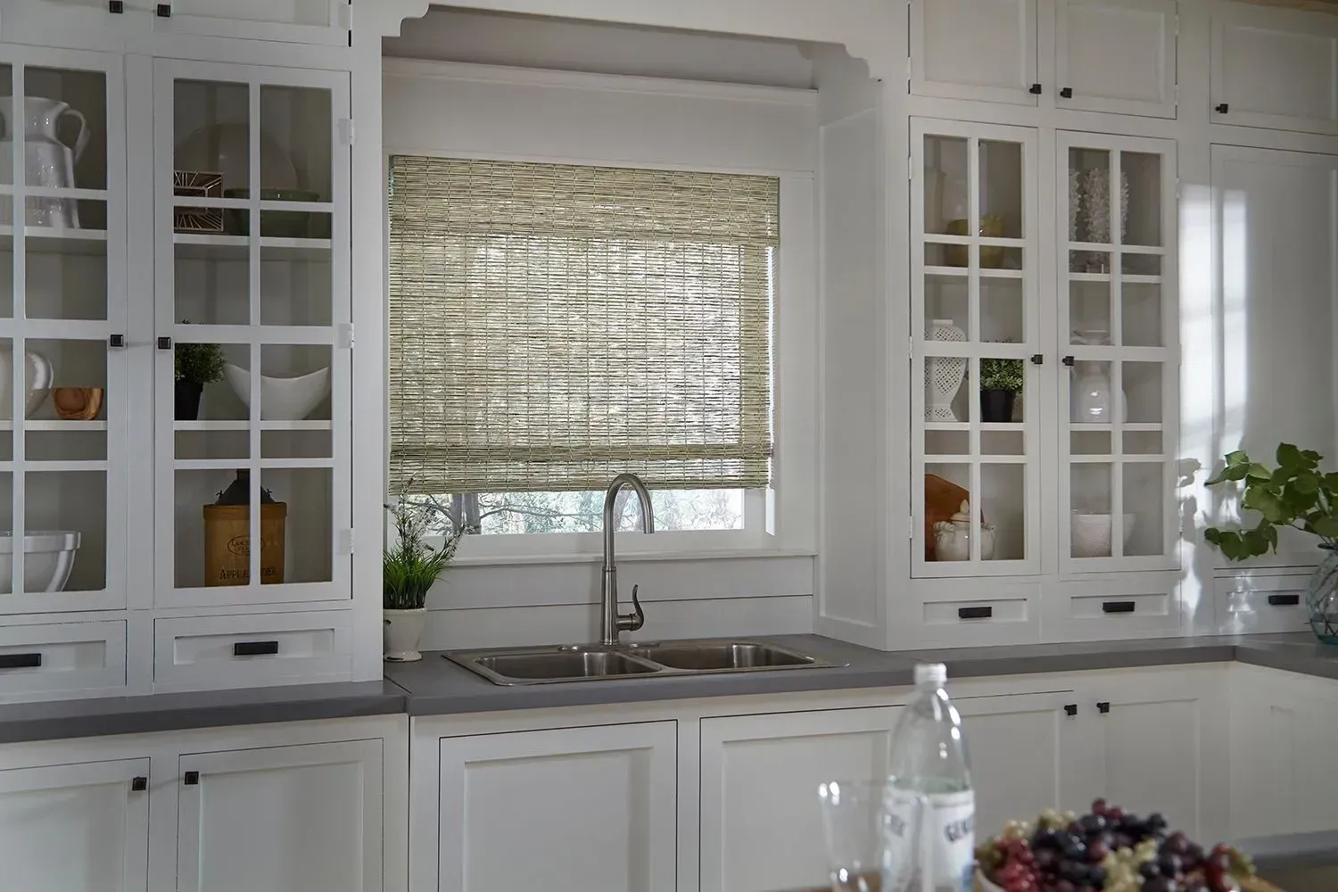 White kitchen cabinets with glass doors flank a window with a woven shade. Stainless steel faucet above a double sink.