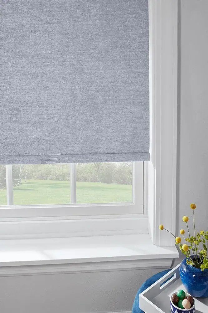 Blue roller blind partially covering a white-framed window, revealing a sunny outdoor scene, flowers on the windowsill.