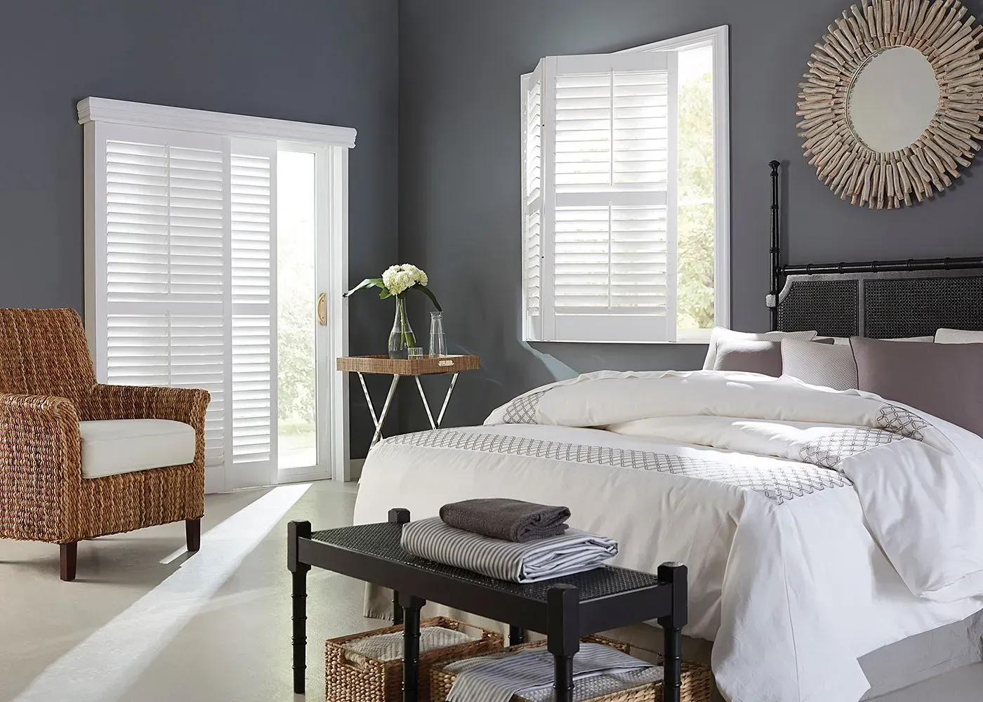 Bedroom with white shutters on a sliding door and window. Bed, wicker chair, and decor present. Gray walls.