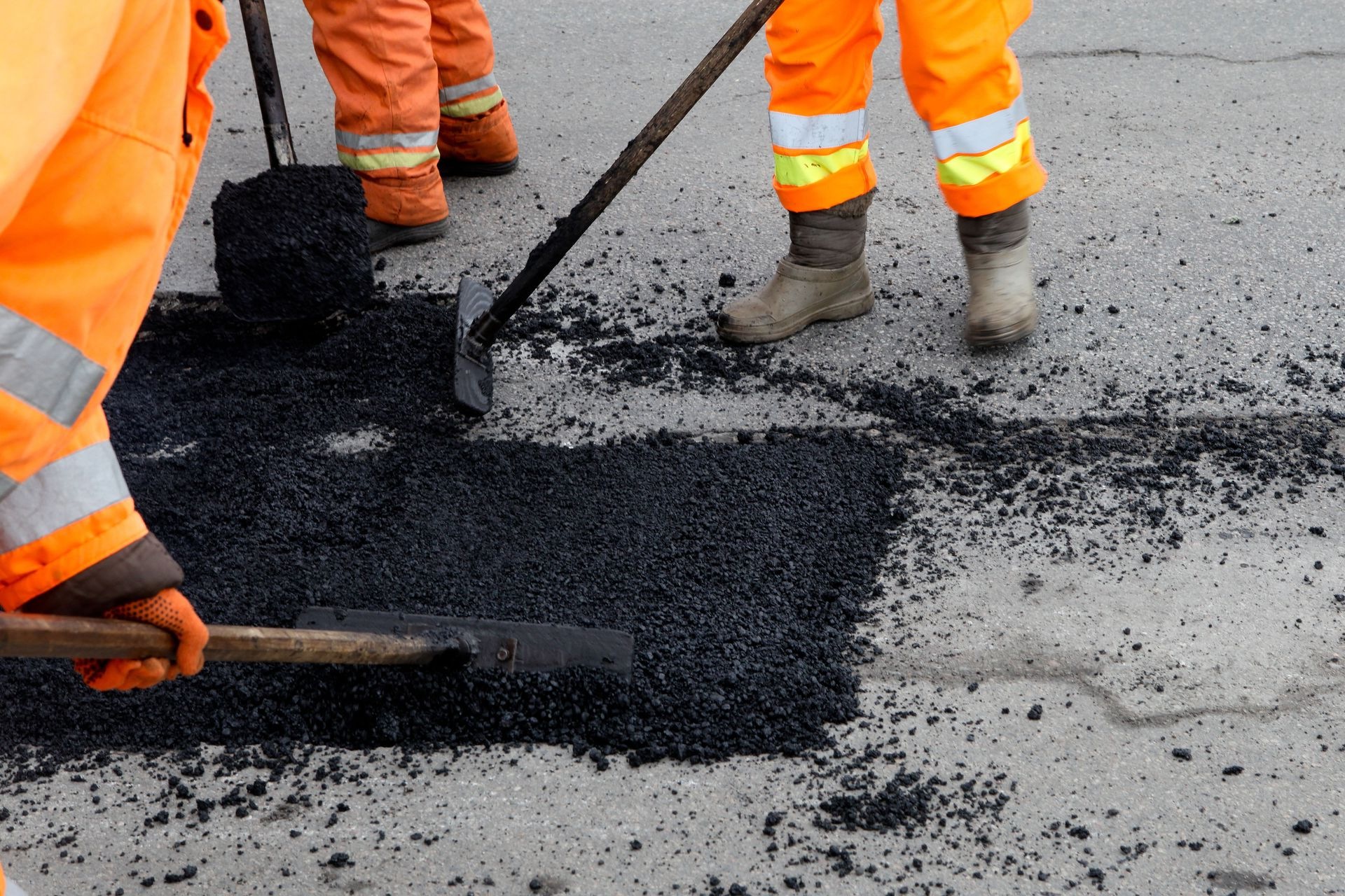 Construction workers in orange high-visibility clothing use shovels and tools to patch a road with fresh asphalt.