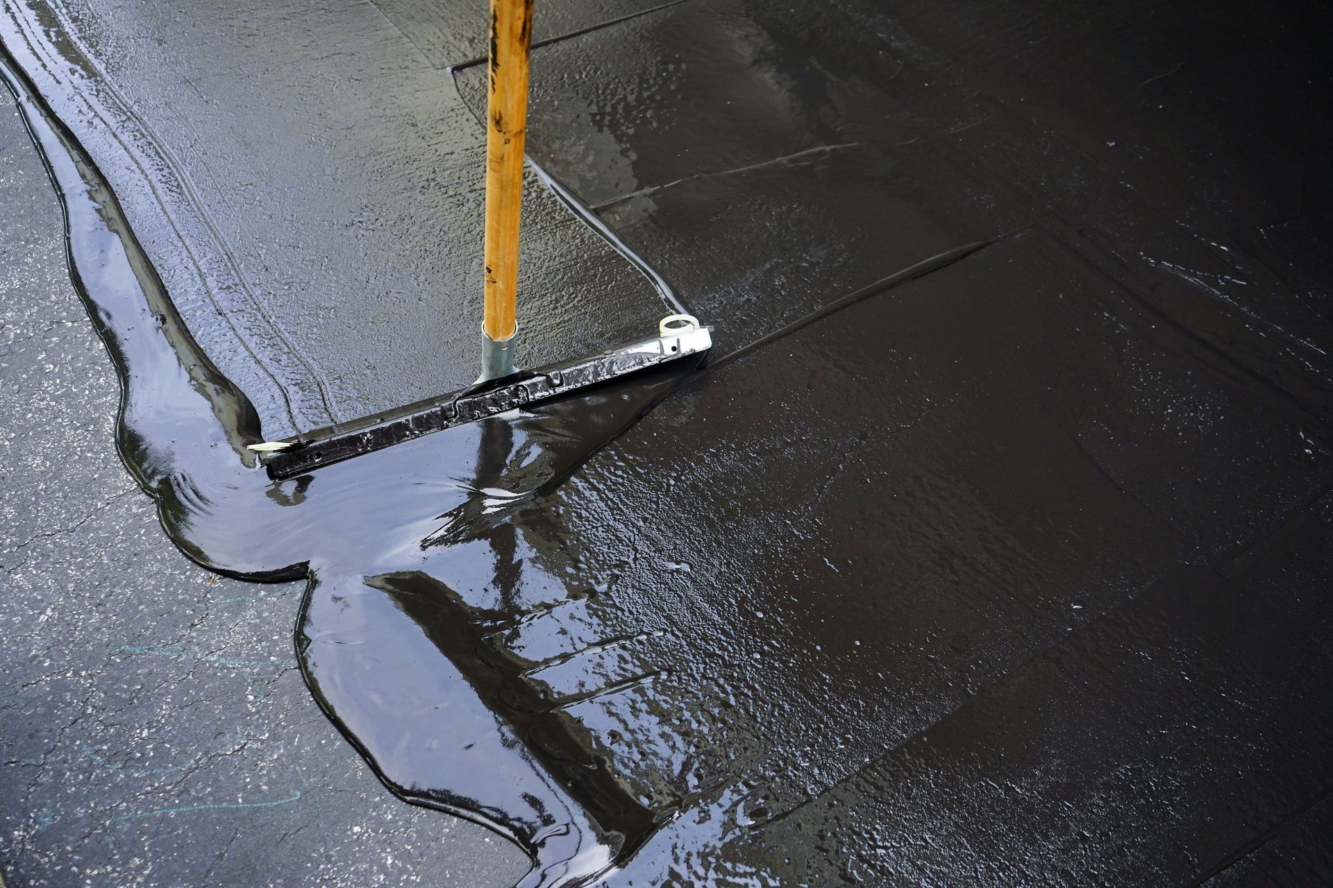 A tool with a wooden handle scrapes dark liquid across a wet, paved surface.