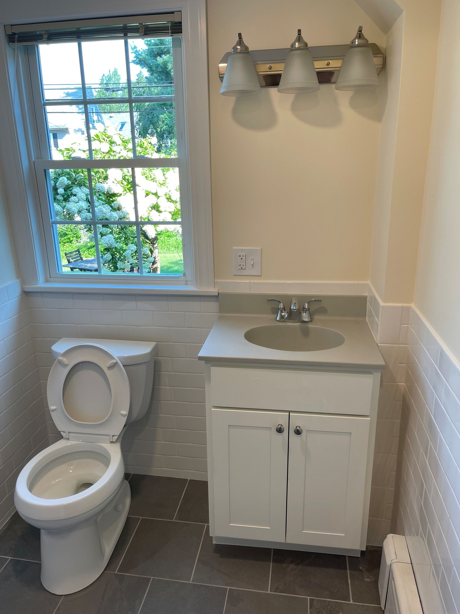 Bathroom with white vanity, toilet, and a window overlooking greenery. Gray tile floor and walls with white textured trim.