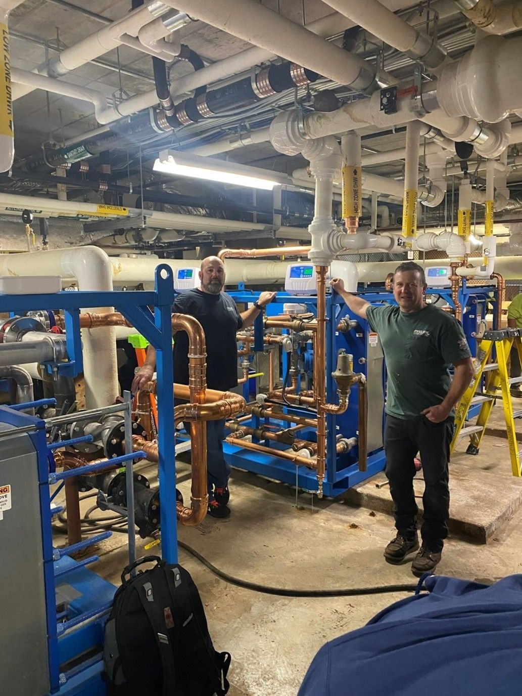 Two men stand beside complex copper piping in an industrial setting.