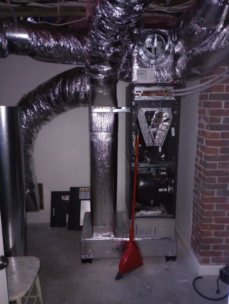 Furnace in basement with ductwork and a red broom. Brick wall, white wall, gray concrete floor.