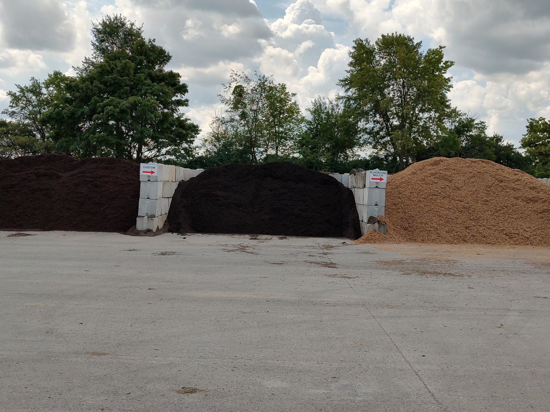 Three piles of mulch: dark brown, black, and light brown, separated by concrete barriers, with trees in the background.