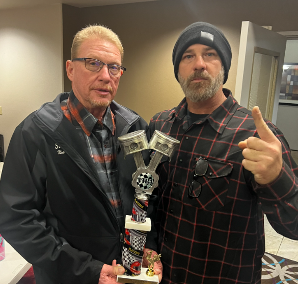 Two men indoors holding a tall trophy, one pointing at the camera; both wearing plaid shirts.
