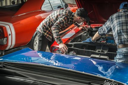 Two mechanics working under the hood of a red car next to a blue car at an auto shop
