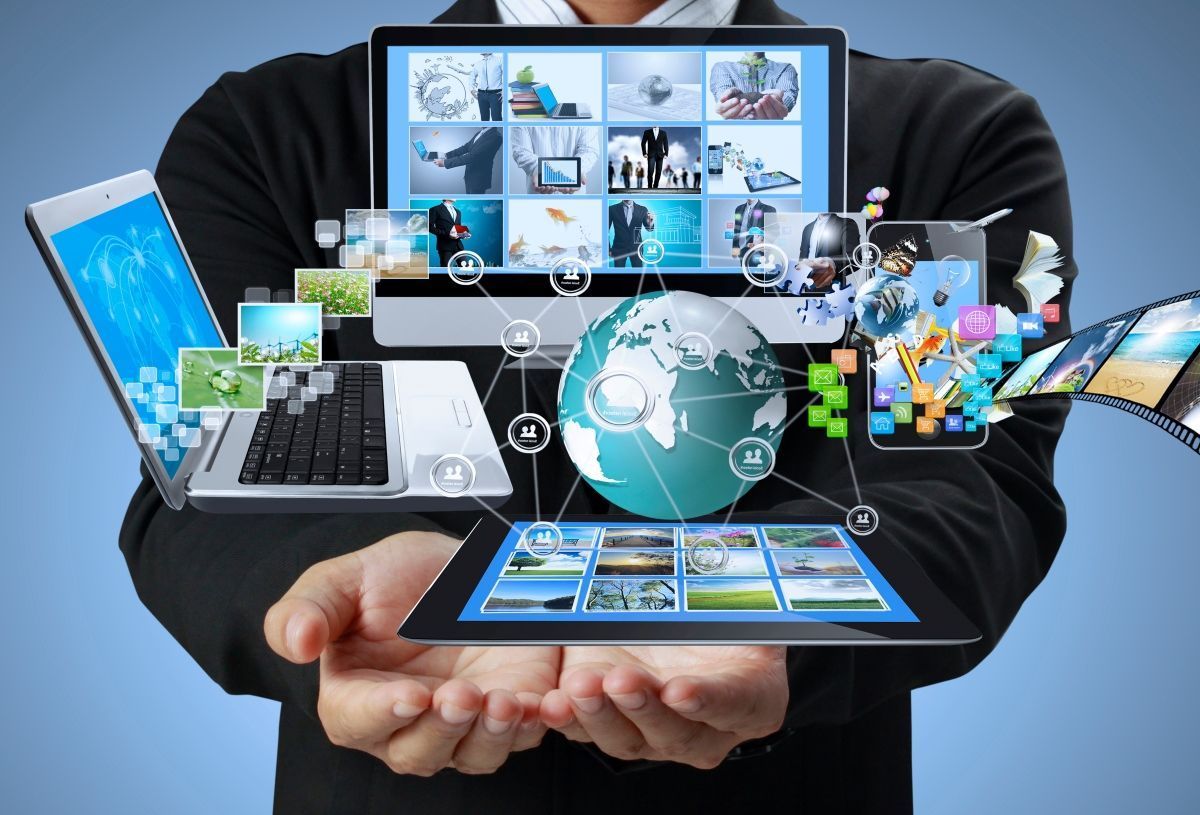 Hands holding a tablet with laptop, globe, and multiple digital device screens showing connected technology icons.