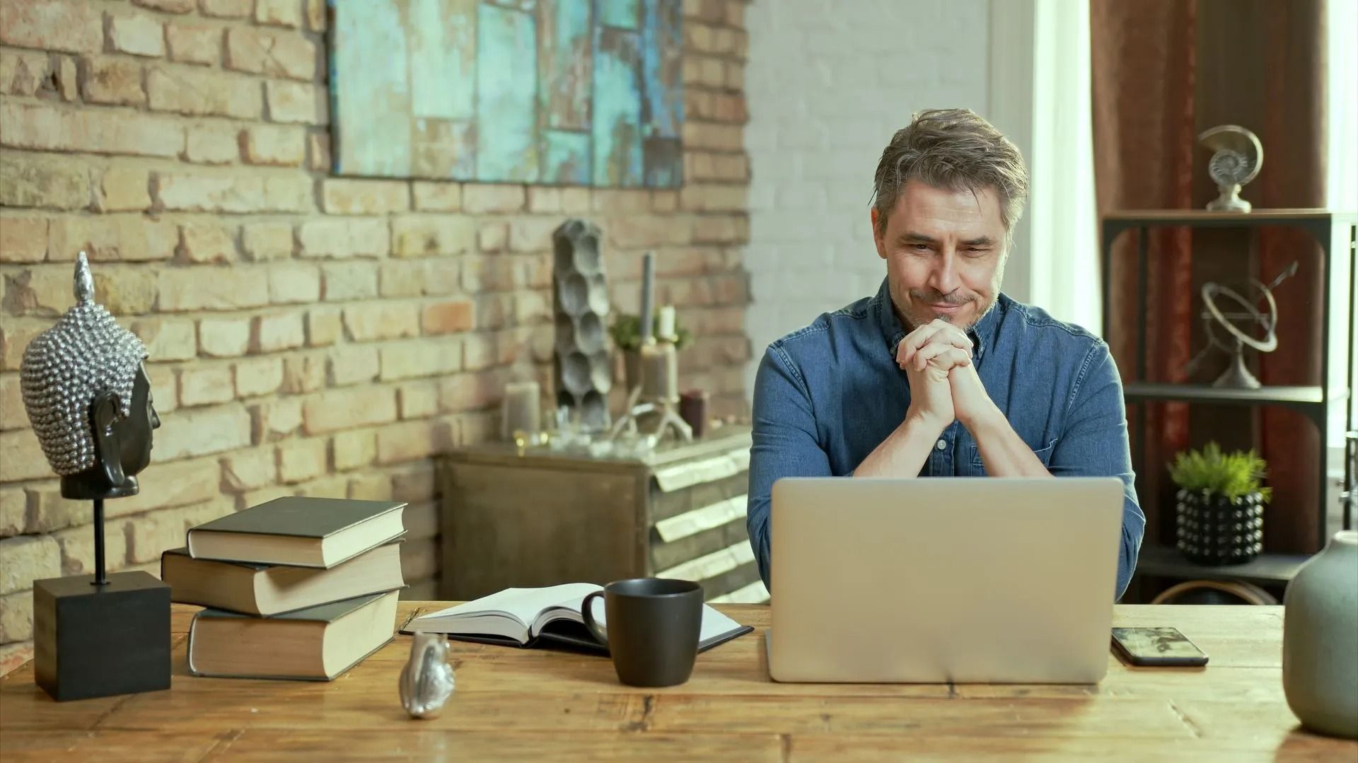 Man seated at a desk working on a laptop in a cozy brick-walled office with books and decor.