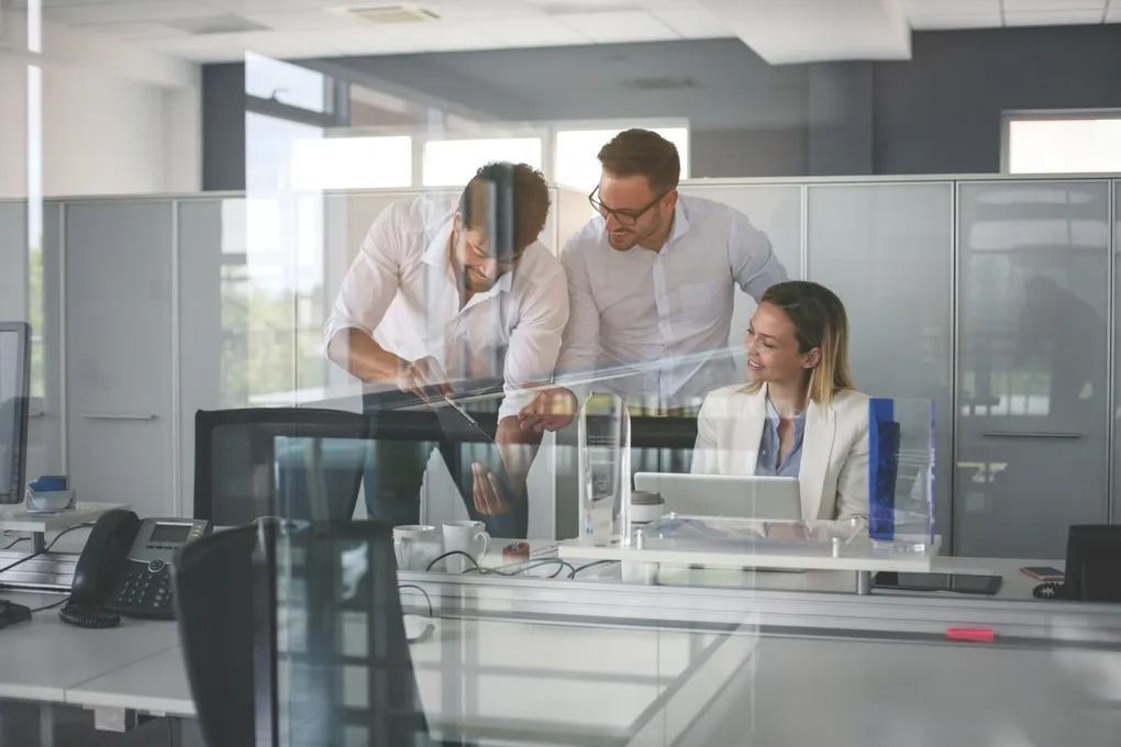 Coworkers collaborating in a modern glass-walled office, reviewing work on a laptop and monitor.