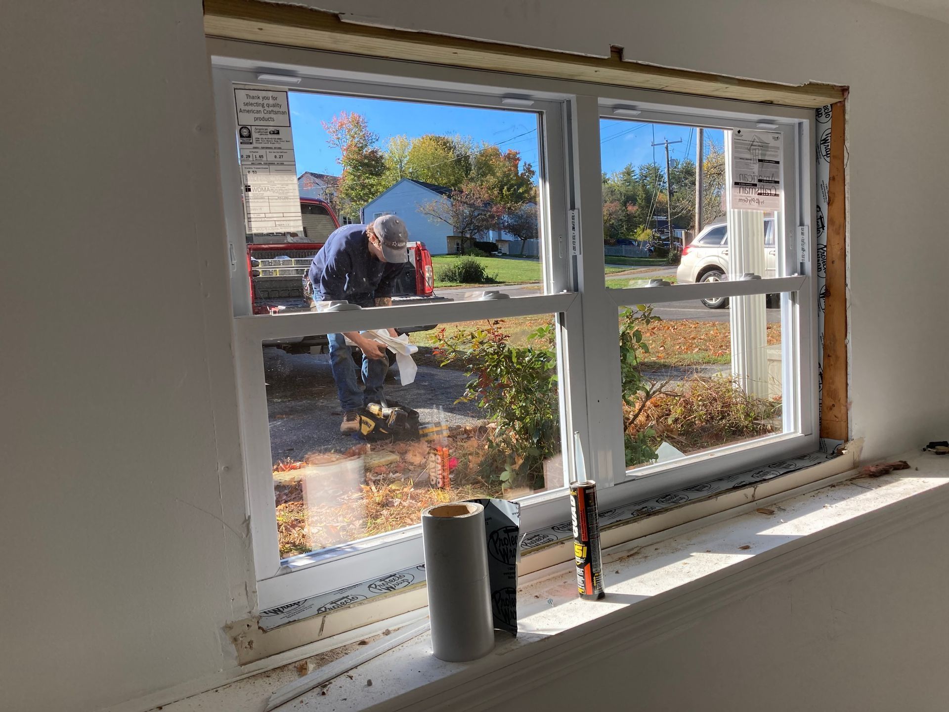 A person installing a white window inside a house; roll of tape and level on the sill.