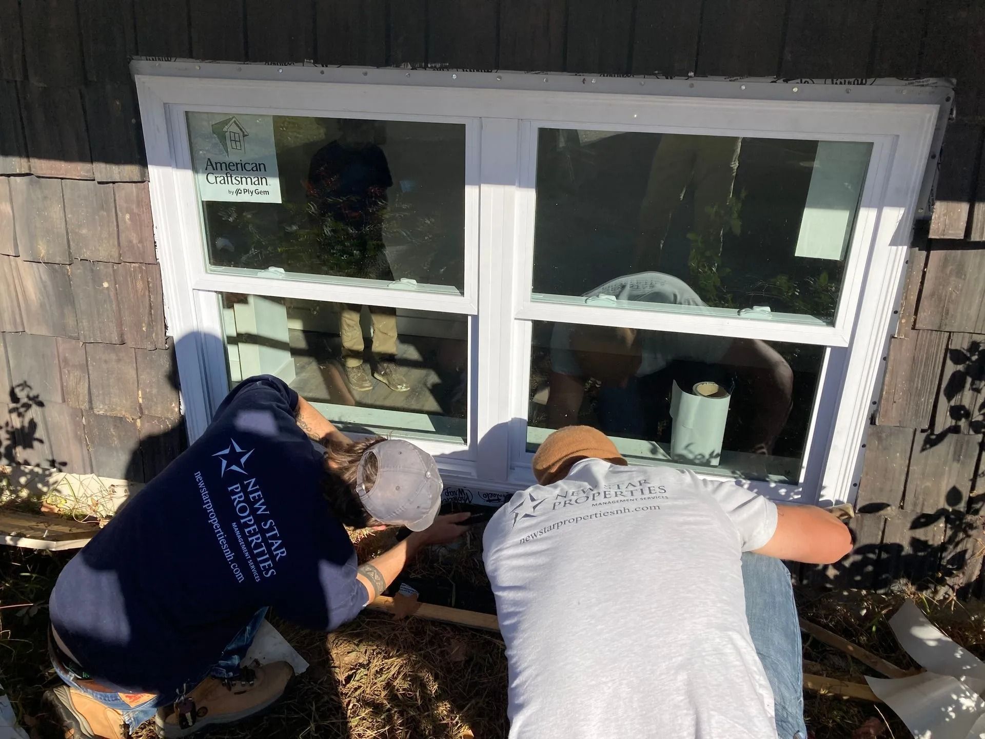 Two people installing a window on a building. They are wearing work clothes, working outside in daylight.