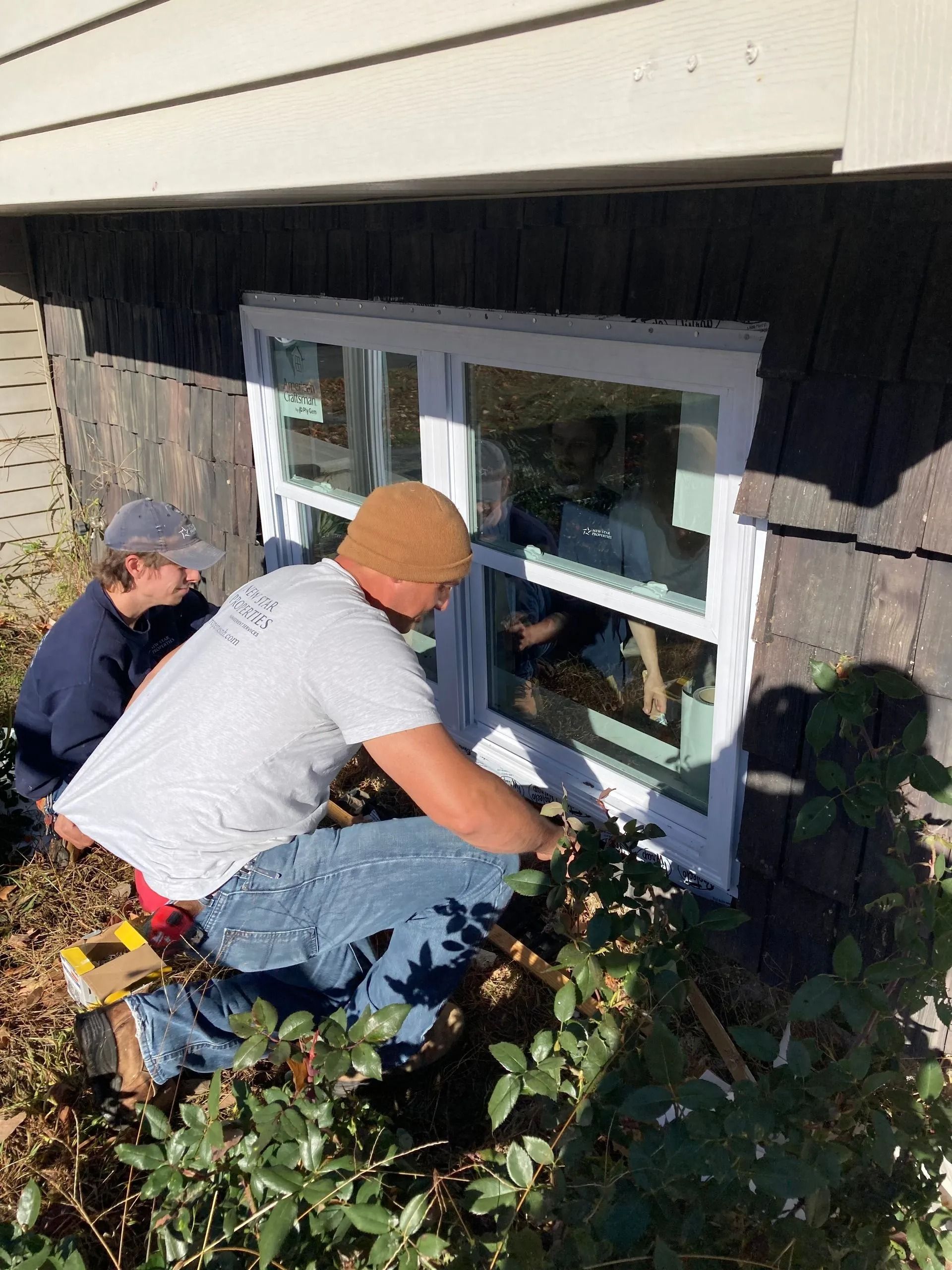 Two people installing a white window in a brown wall. One person is kneeling, applying something to the window frame.