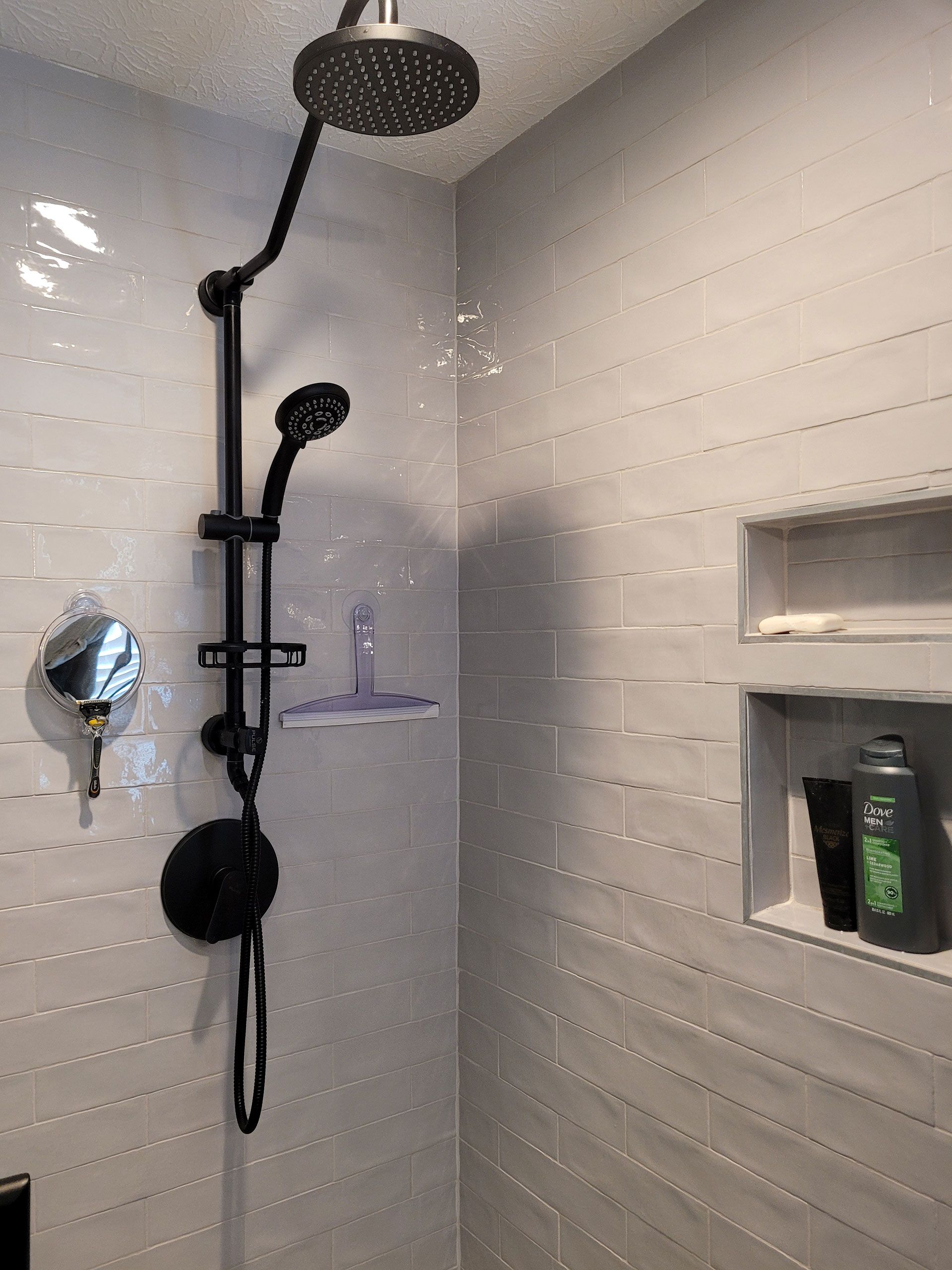 Black shower fixtures against gray tiled walls with built-in shelf containing toiletries.