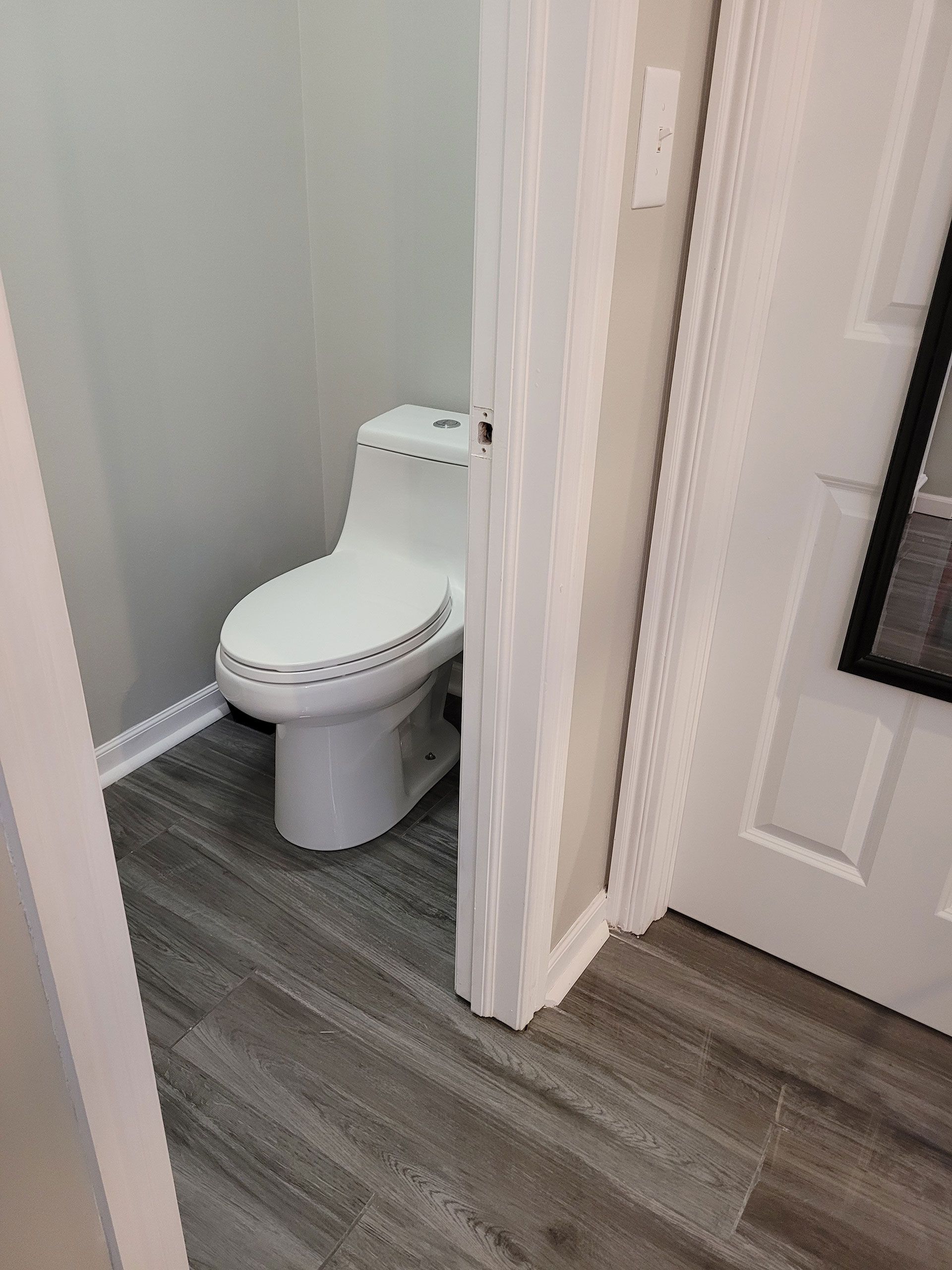 White toilet in a small bathroom with gray walls and wood-look flooring. Partially visible white door and trim.