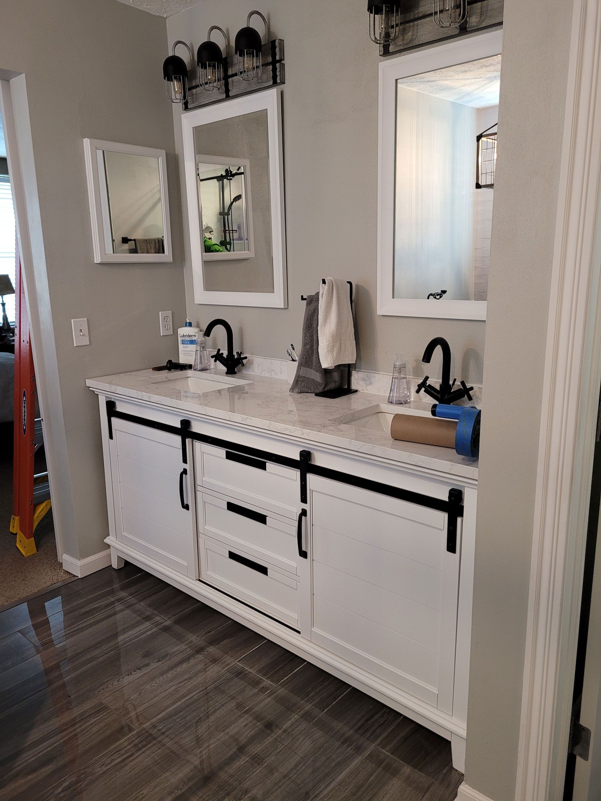 White bathroom vanity with two sinks, black faucets and hardware. Gray walls, mirrors, and lighting.