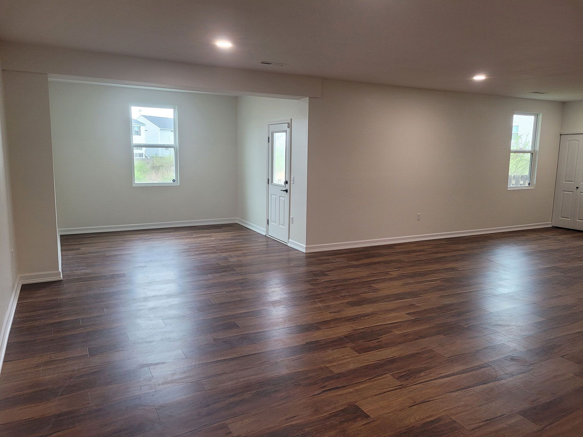 Empty room with hardwood floors, white walls, and a doorway. Two windows and recessed lighting visible.