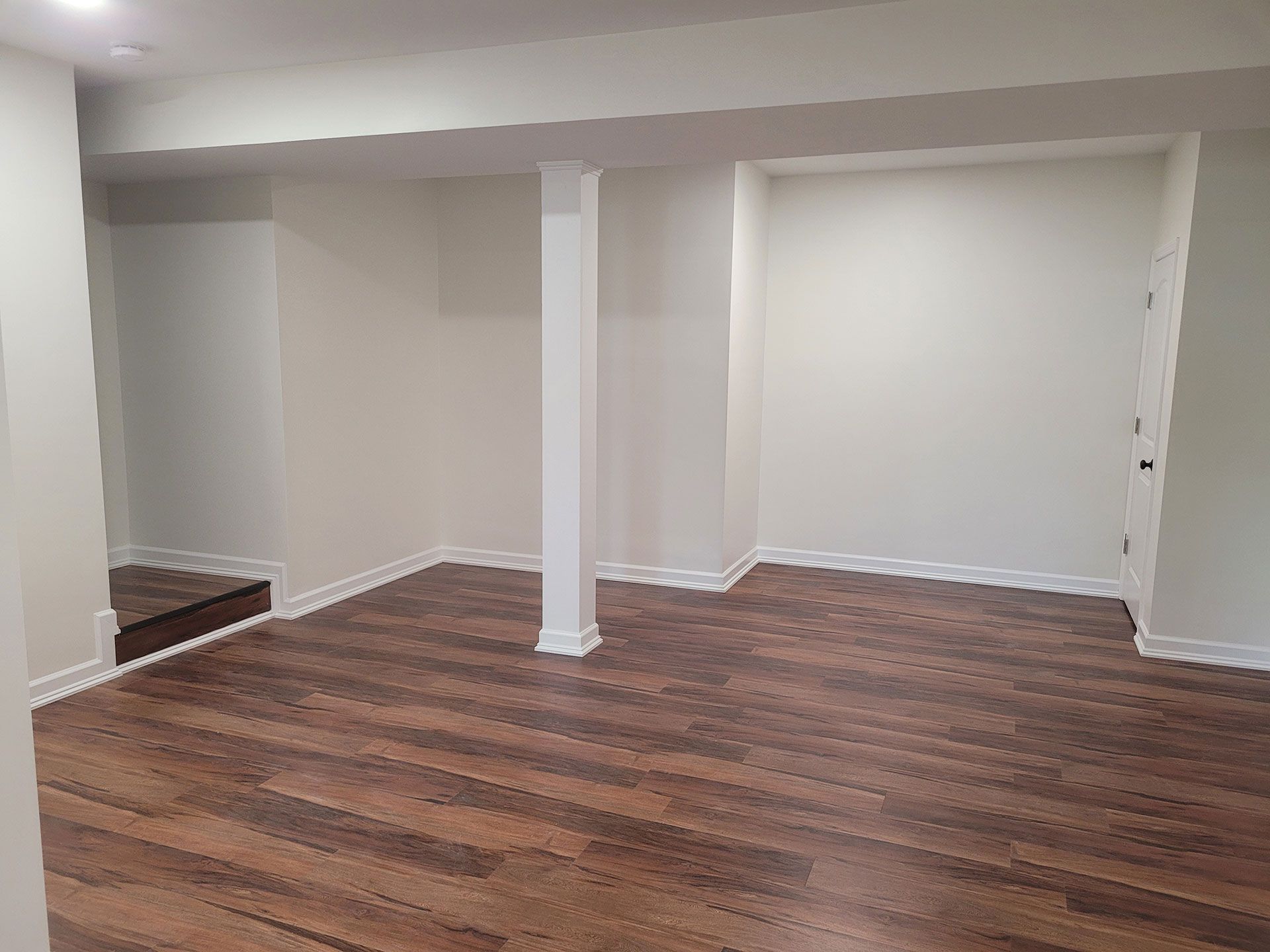 Empty, freshly painted basement room with wood-look flooring, a support pillar, and a recessed wall area.