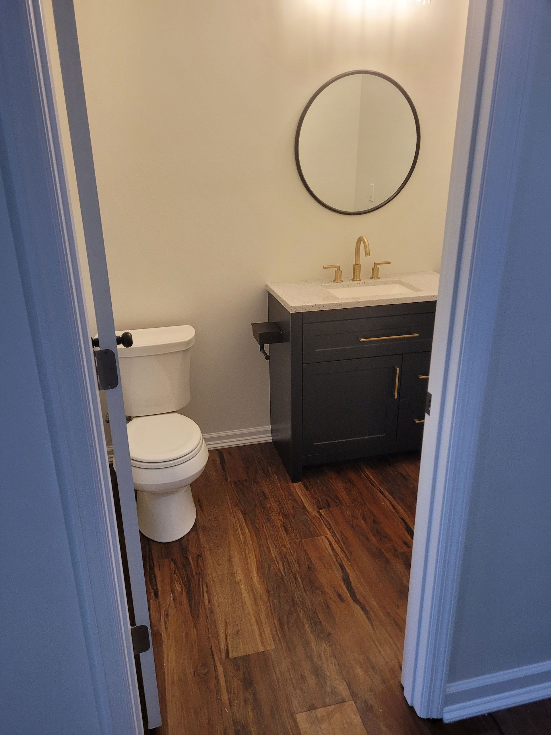 Small bathroom with dark vanity, round mirror, and wood-look flooring.