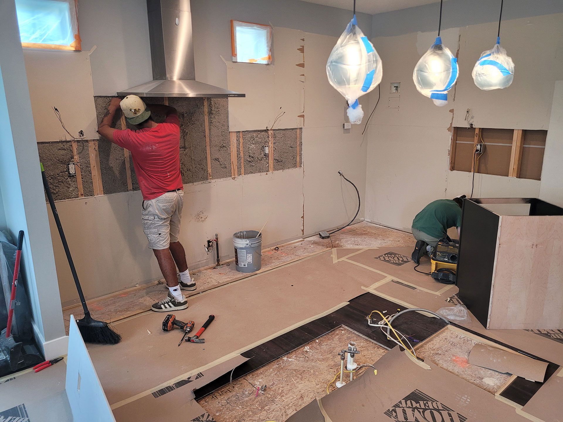 Kitchen remodel: Two workers install flooring and range hood in a room with exposed walls and electrical wiring.