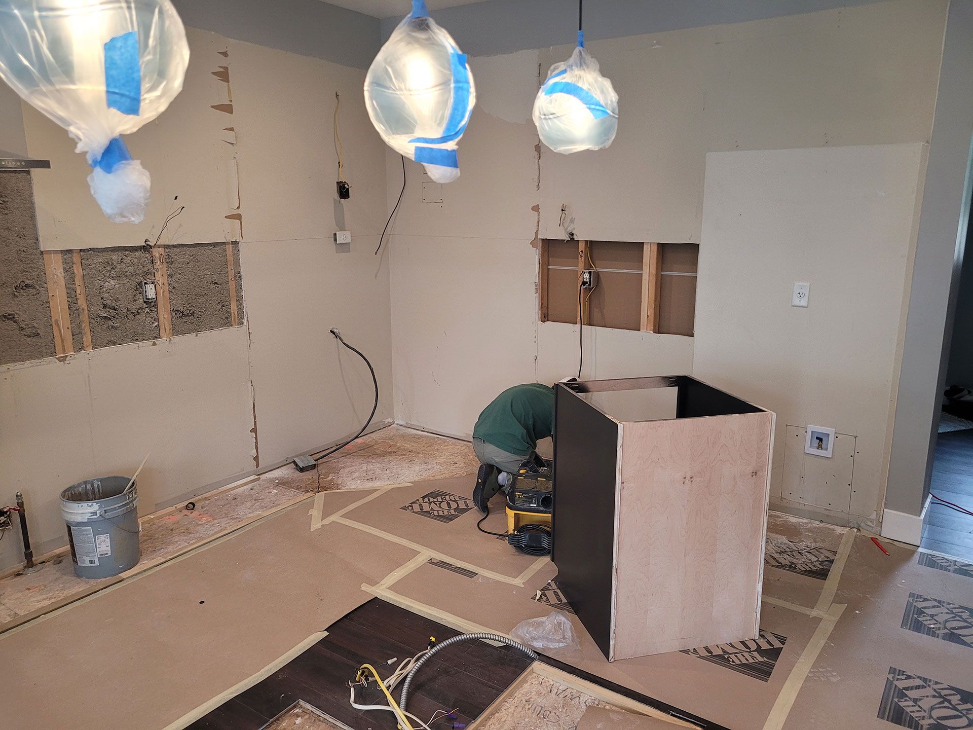 Kitchen under construction: worker, tools, exposed walls, plywood, water-filled bags hanging, floor markings.