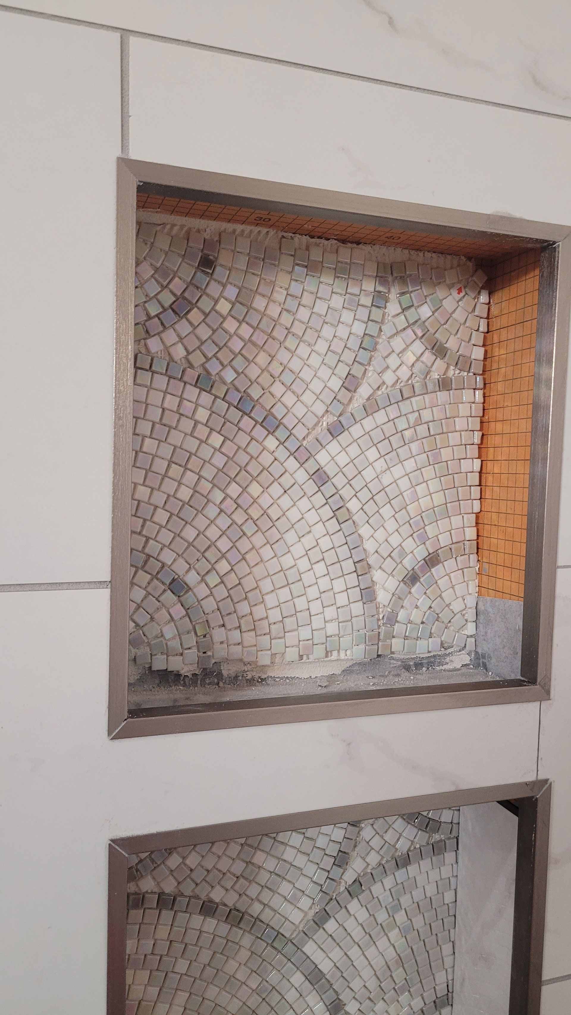 Tile niche with mosaic tile design, framed in silver, set in white tiled wall.
