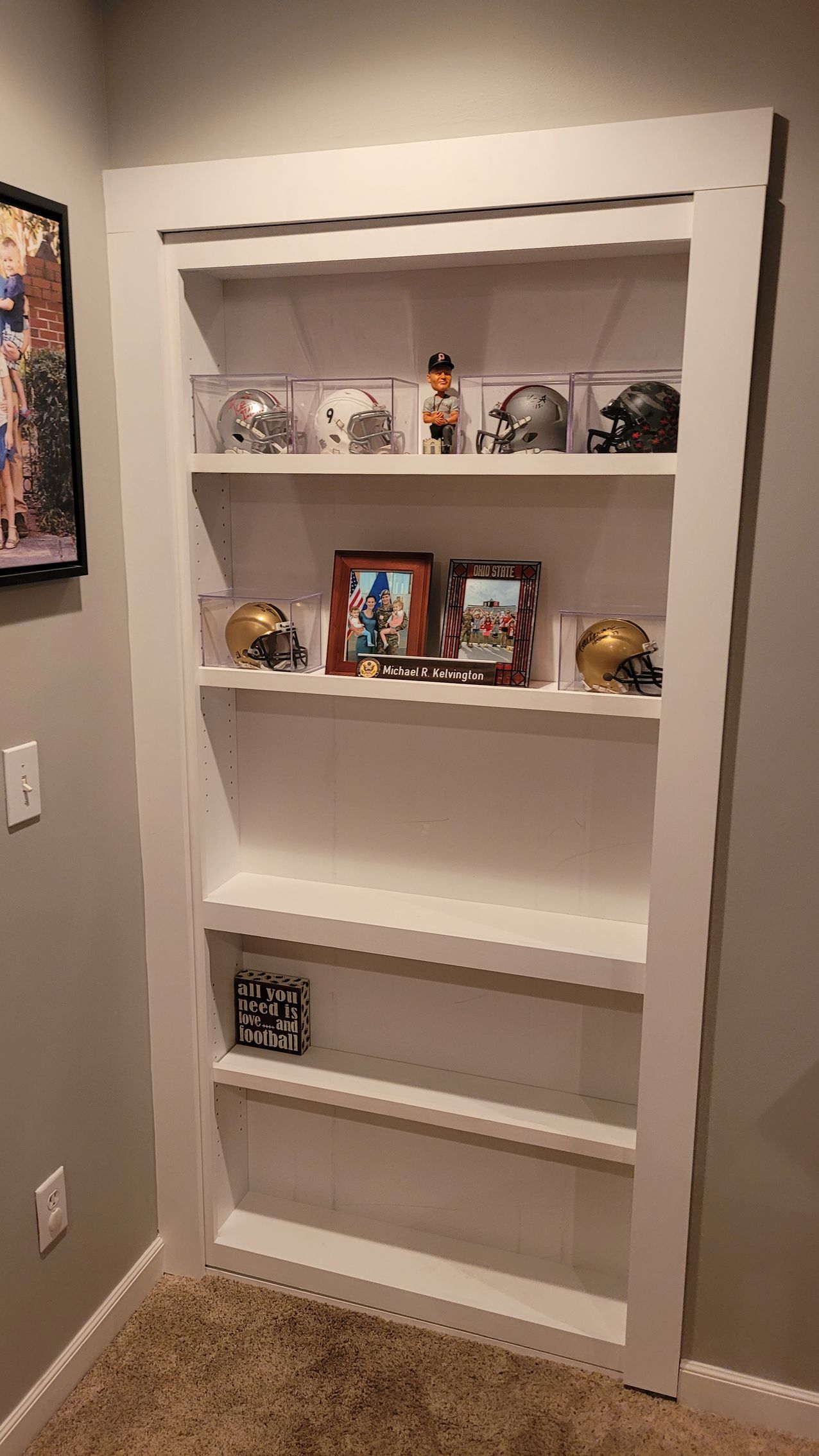 White built-in bookshelf in a wall alcove displays football helmets, framed photos, and other collectibles.