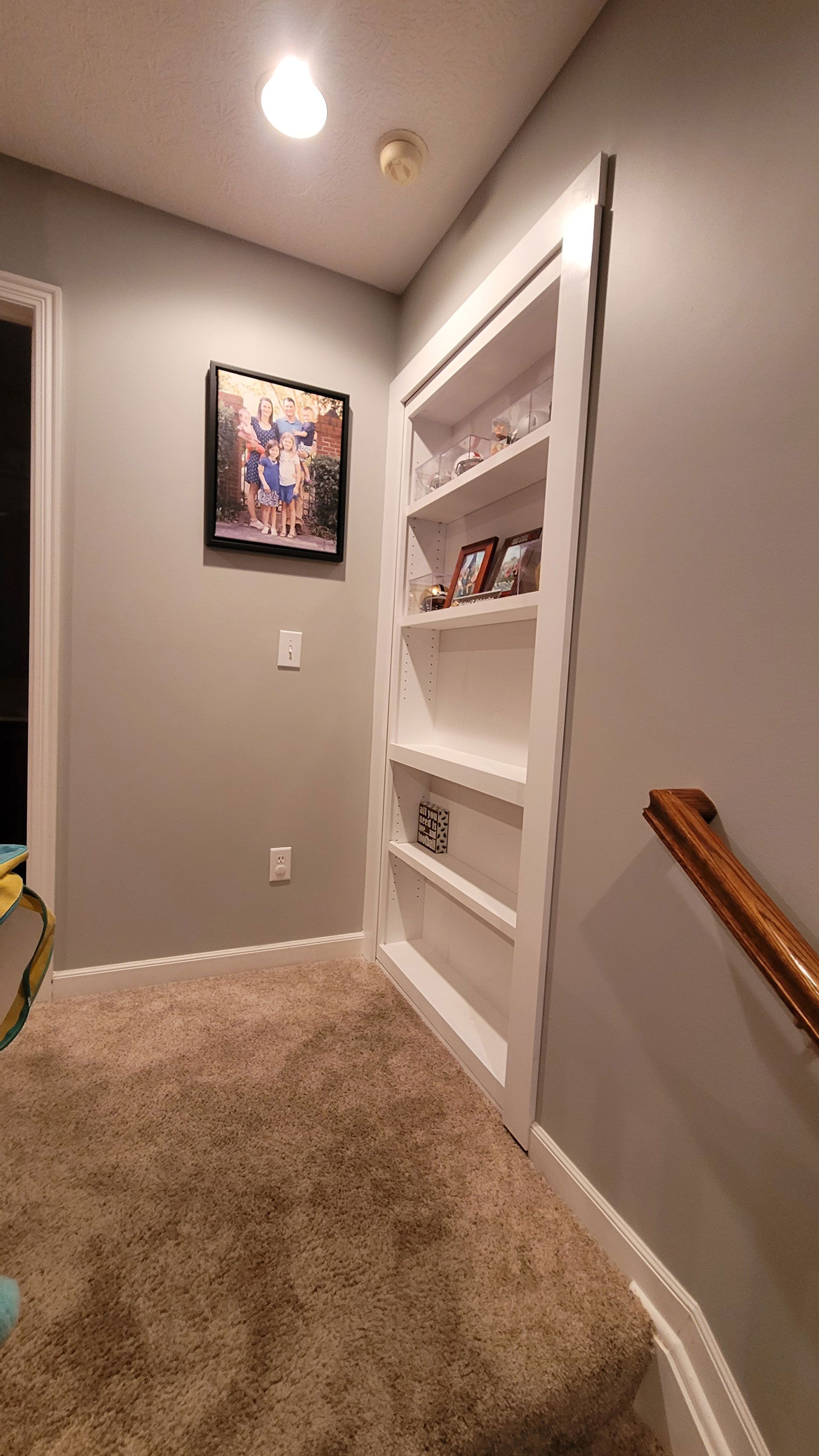 A built-in white bookshelf in a gray hallway, next to a staircase with a wooden handrail.