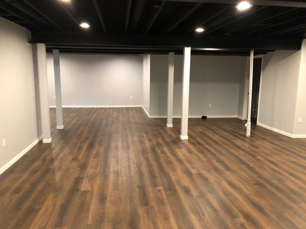 Empty renovated basement with wood-look flooring, gray walls, black ceiling, and white support columns.