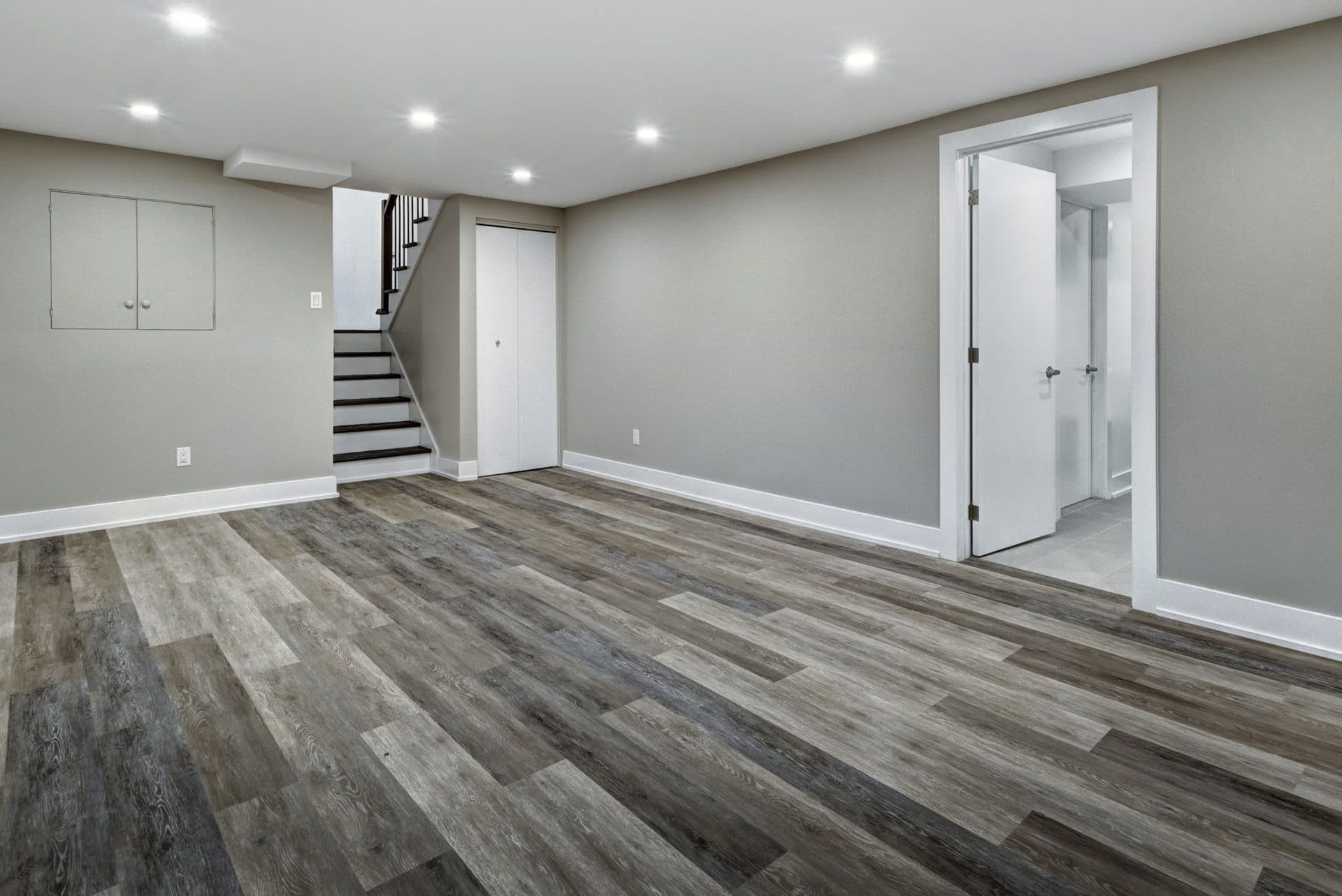 Empty basement room with gray walls, wood-look floor, doors, and staircase.
