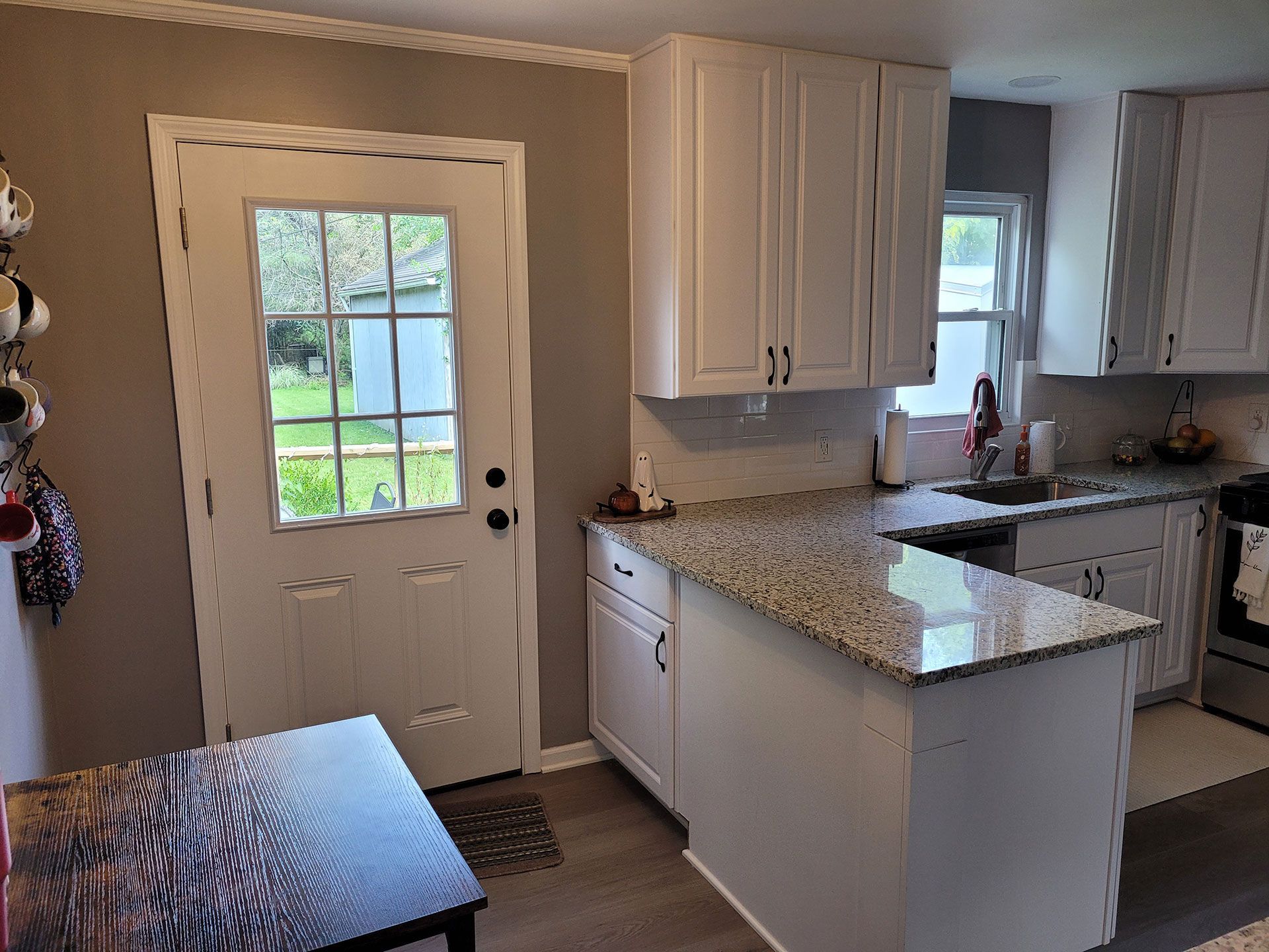 White kitchen with island, cabinets, door, granite countertops. Dark table and mug rack on wall.