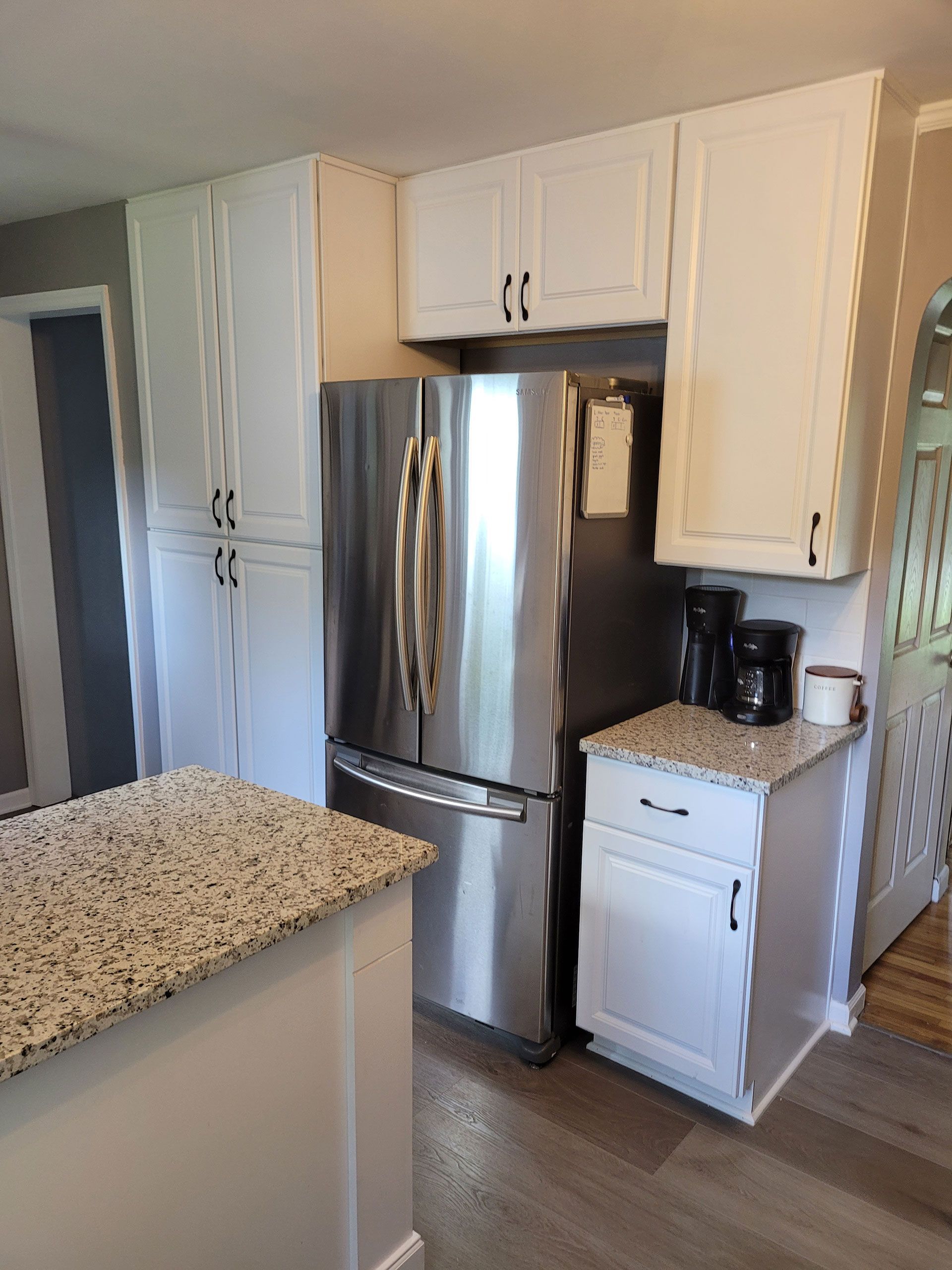 White kitchen cabinets surround a stainless steel refrigerator. Granite countertop, light wood floor.