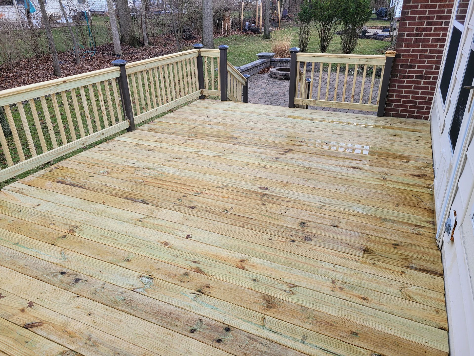 Wooden deck with railings. Steps lead to a yard. The deck is attached to a brick wall.